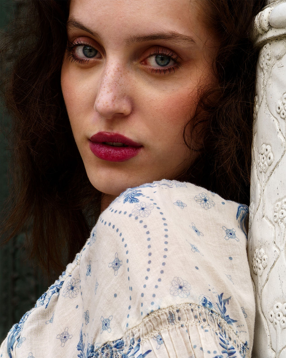 Vertical Bucchianeri editorial fashion portrait of a woman in a cream-toned floral dress looking over her shoulder beside a classical stone column.