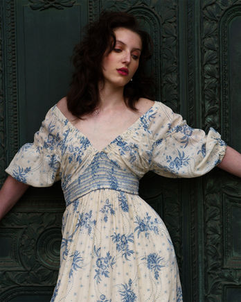 Outstretched arms and a quiet downward gaze define a woman in a puff-sleeve ivory dress against green patina doors.