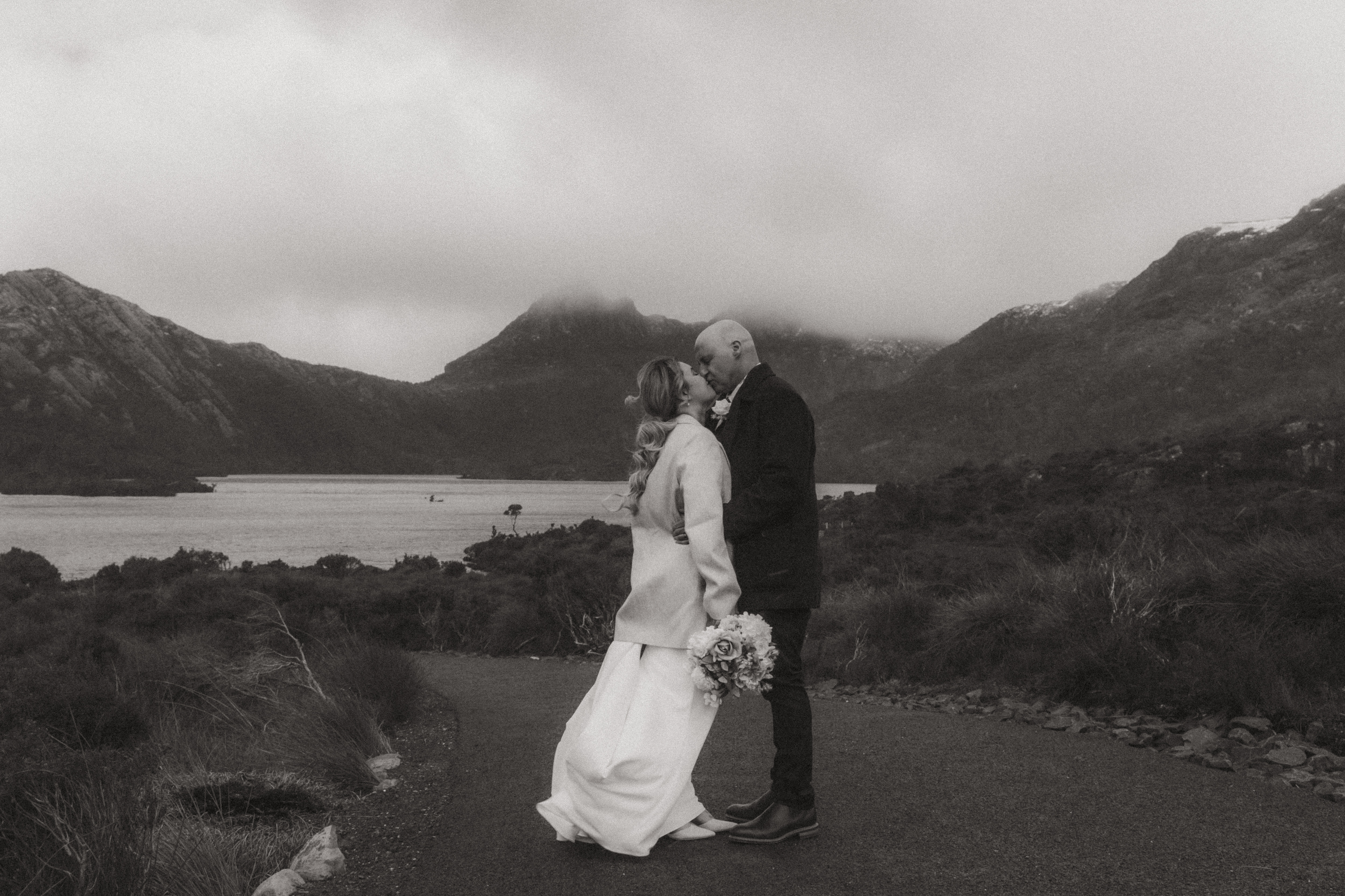 A black and white frame of a bride in a long coat and gown kissing a groom on a sealed path beside a lake, a cloud capped peak sitting between two ridges behind them and low scrub on either side.