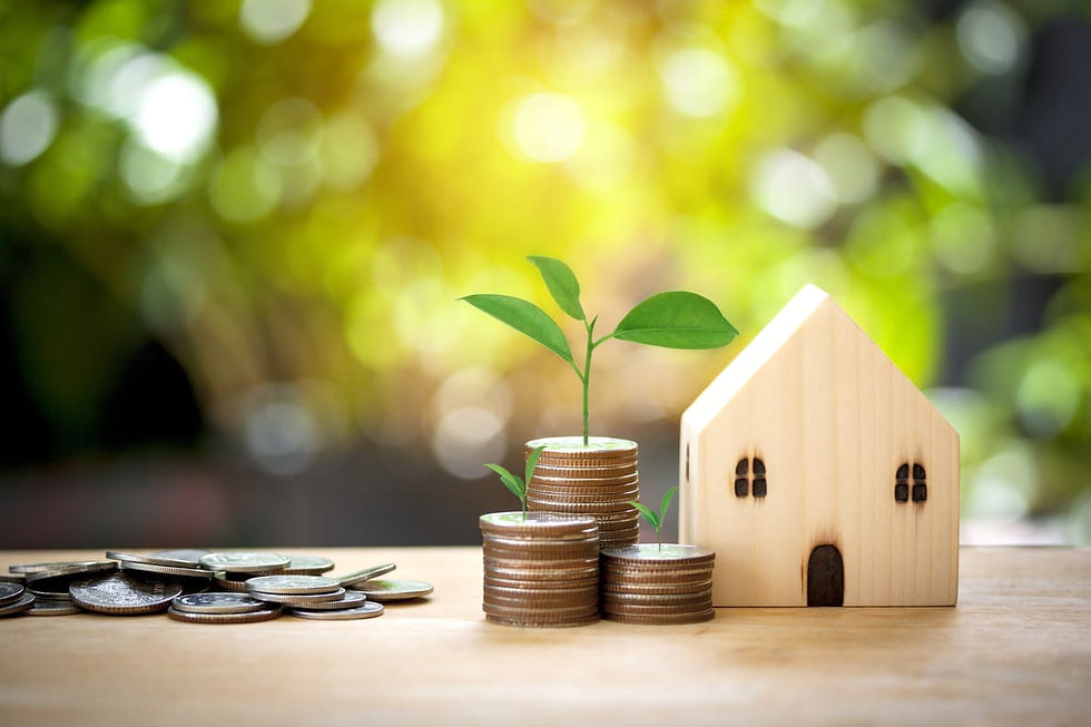 A small wooden house beside stacks of coins with sprouting plants, on a wooden surface. Blurred green background suggests growth and investment.