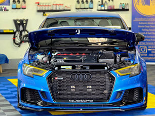 Blue Audi with an open hood in a garage. Focus on engine details, yellow headlights, and "quattro" text on grille. Tools hang on walls.