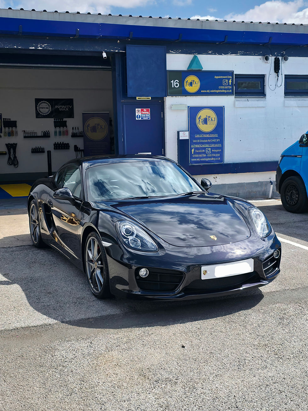 Shiny black sports car parked outside a detailing service, with blue and white signs, under a partly cloudy sky.