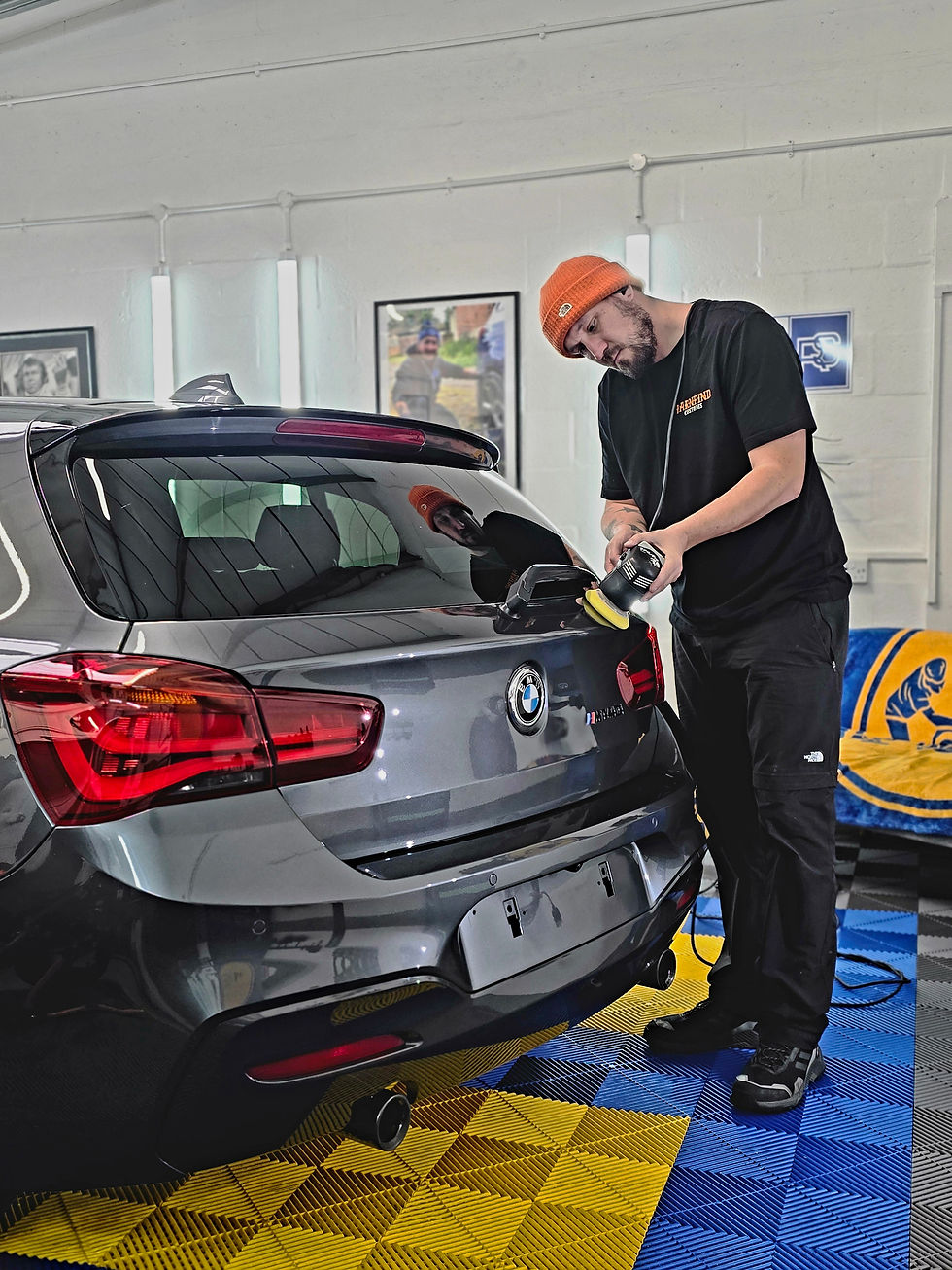 Man polishing a grey BMW in a garage. He's wearing an orange beanie and black attire. Blue and yellow patterned floor.