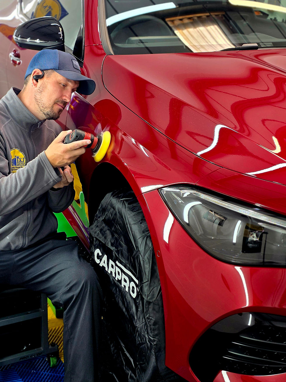 Man in cap polishes a red car’s fender with a buffer in a garage; black wheel cover reads CARPRO.