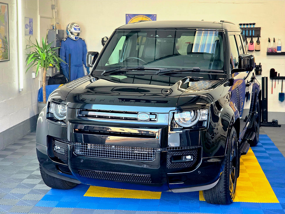 Black Land Rover Defender in a garage with checkered blue and yellow floor. Racing suit and helmet hang in back; detailing tools are visible.