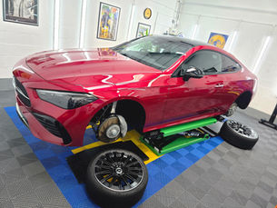 Red Mercedes coupe on a lift in a workshop, wheels removed and spare rims on the floor, under bright lights.