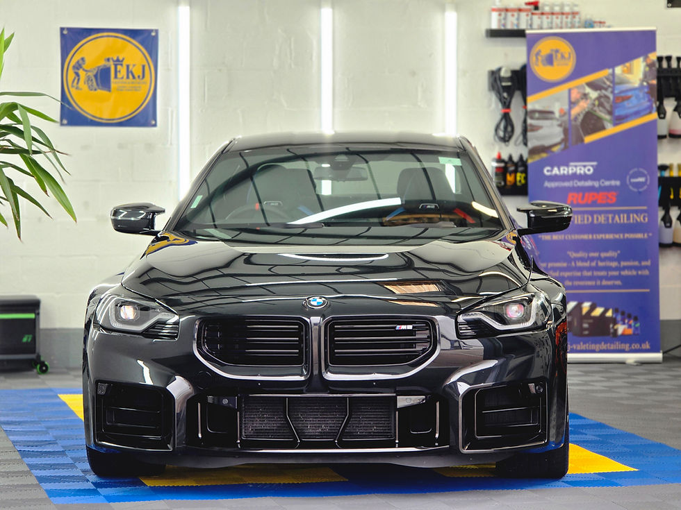 Black BMW M2 in a car detailing shop, with CARPRO and RUPES banners and bright reflections on its glossy hood