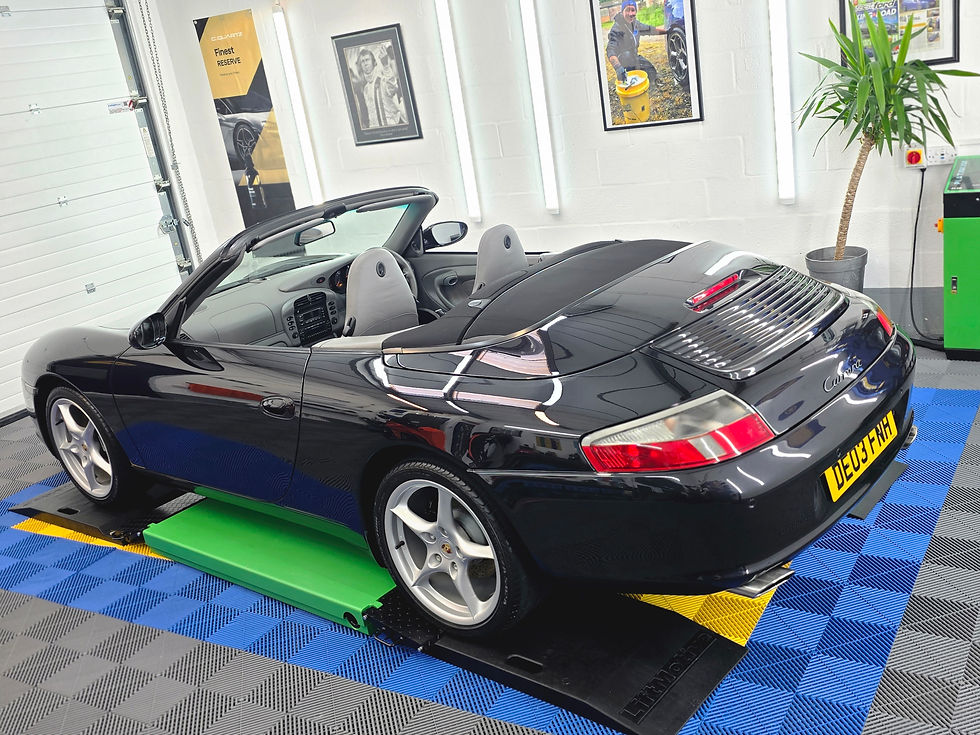 Black Porsche Cabriolet on a blue-and-gray showroom floor, with wall posters, a potted plant, and a yellow license plate.