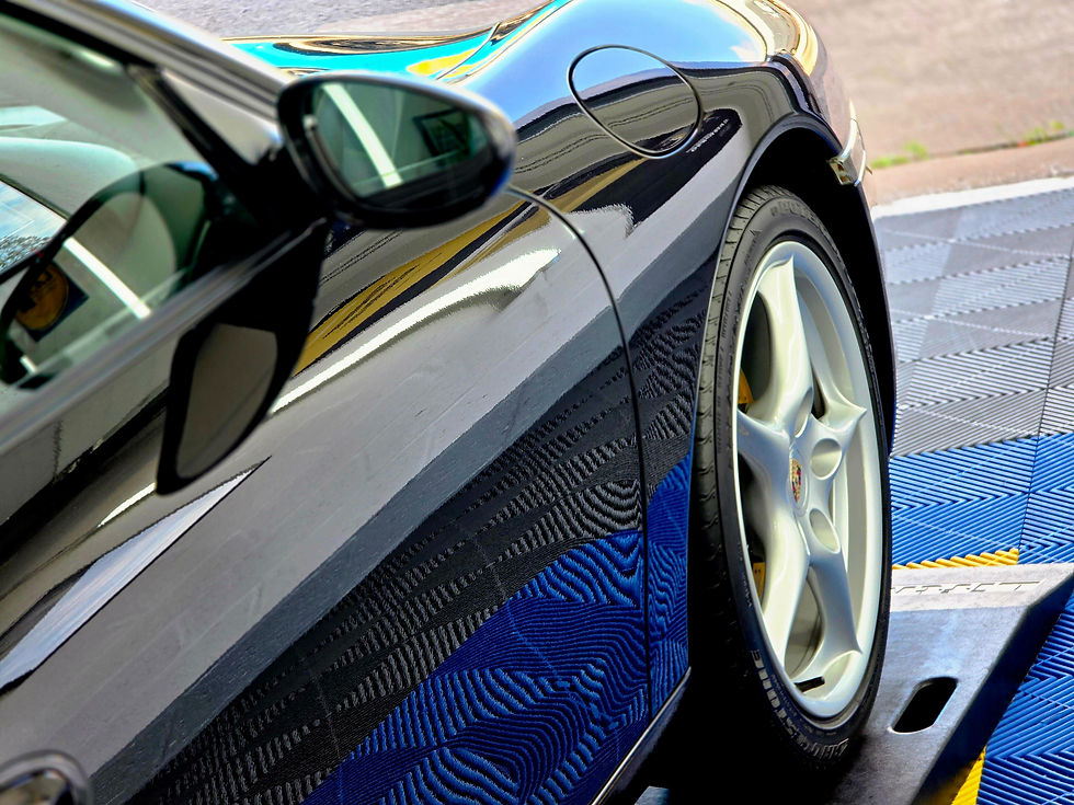 Close-up of a glossy black Porsche sports car on a blue service platform, showing mirror, fuel cap, and wheel reflections.