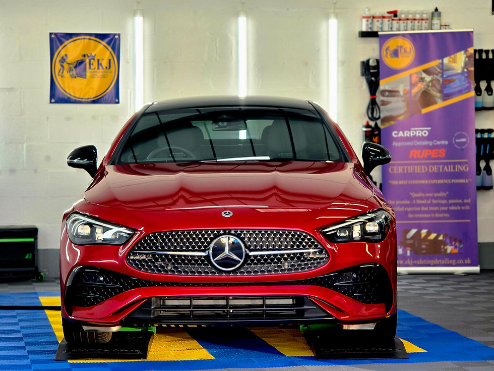 Red Mercedes sedan in a detailing shop, flanked by EKJ and CARPRO/RUPES banners, on blue-yellow floor mats.
