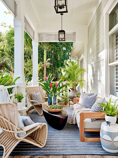 Front porch seating area with woven chairs, layered cushions, sculptural planters, lantern lighting, and lush greenery.
