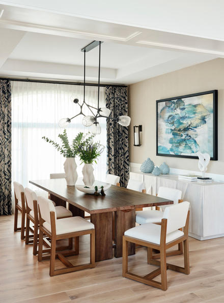 Dining room with large wood table, upholstered chairs, sculptural pendant lighting, soft drapery, and curated artwork.