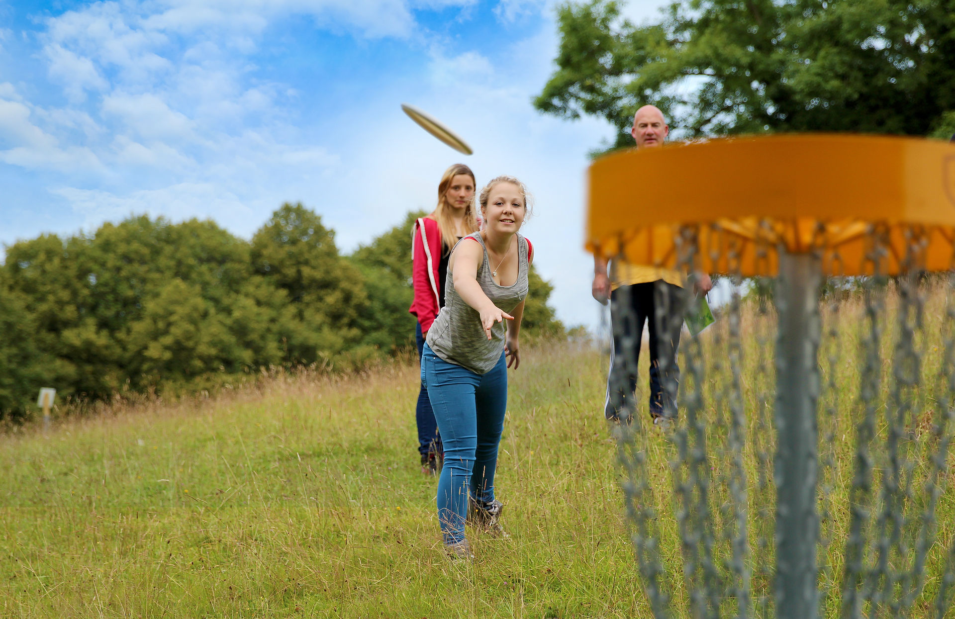 A woman has thrown a frisbee towards a frisbee golf basket. The disc is in mid-air, floating towards the hanging chains of the basket. Trees and blue skies fill the background.