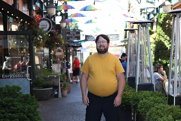 Photo of Jamie Wellbourn, man with beard wearing glasses, in yellow t-shirt with black shorts in alley in Sidney with brick path, green potted plants, shop fronts, restaurant patio, and rainbow umbrellas hanging above