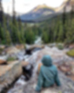 Young female hiker sits atop a rock beside flowing evergreen forest stream surrounded by high mountain peaks