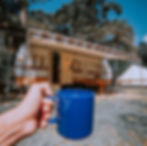 Young person holding up blue mug next to G40 RV camper with awning setup in the forest