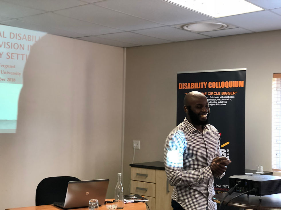 Man with hands clasped speaking to an implied audience with a banner behind him that says Disability Colloquium