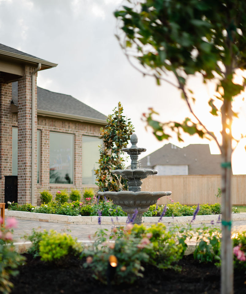 Three-tier garden fountain surrounded by elevated stone flower beds, blooming shrubs, and trees in a landscaped backyard at sunset.