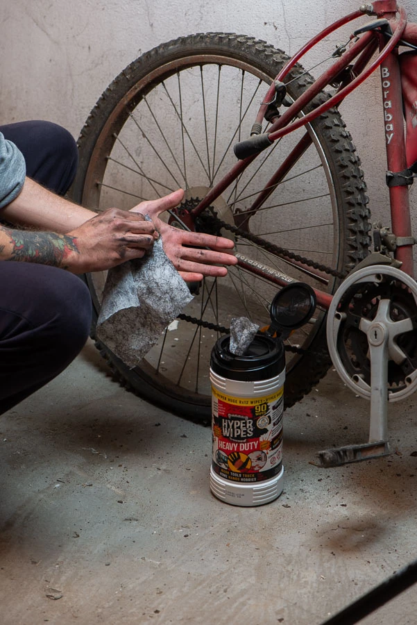 Worker wiping dirty hands beside a bicycle and an open Hyper Wipes 90-count canister