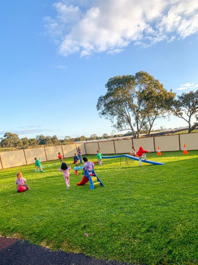 A wide view of a large green grass outdoor playground at Clementson Drive Early Educational Centre under a bright blue sky. Children are enjoying active play across the lawn, including riding hopper balls, climbing a small plastic slide, and walking up a blue balance ramp. Demonstrates the extensive physical play space available for families near Leppington, Kemps Creek, Austral, and Bradfield.