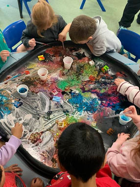 Overhead view of a group of young children gathered around a large black sensory tray filled with colorful glitter, liquids, foam, and letter blocks. The children are using plastic pipettes for hands-on fine motor and sensory experimentation at Clementson Drive Early Educational Centre, serving families near Leppington, Kemps Creek, Austral, and Bradfield.
