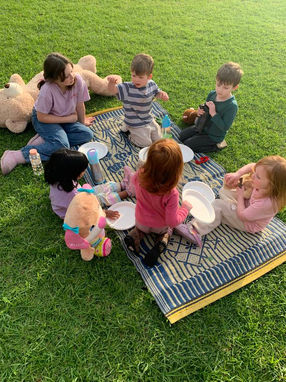 A group of six young children sitting in a circle on a patterned blue woven mat on green grass, enjoying an outdoor teddy bear picnic. The children have white paper plates, water bottles, and plush stuffed animals, including a giant teddy bear resting nearby. Demonstrates outdoor social connection and play-based learning at Clementson Drive Early Educational Centre, serving Leppington, Kemps Creek, Austral, and Bradfield.