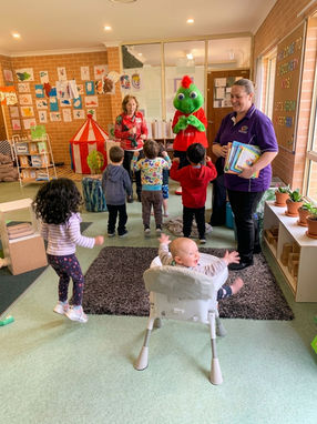 Young children and early childhood educators happily dancing and jumping during a live interactive performance with a green mascot character inside a bright classroom. In the foreground, an infant sits smiling in a high chair with arms raised. Illustrates dynamic music and movement programs at Clementson Drive Early Educational Centre, serving Leppington, Kemps Creek, Austral, and Bradfield.
