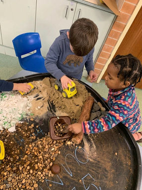 Two young children engaged in sensory play around a large black tray filled with kinetic sand, pebbles, small logs, and yellow toy construction trucks. A boy pushes a dump truck through the sand while a girl uses a small scoop to gather gravel, illustrating fine motor and STEM-focused learning at Clementson Drive Early Educational Centre, serving Leppington, Kemps Creek, Austral, and Bradfield.