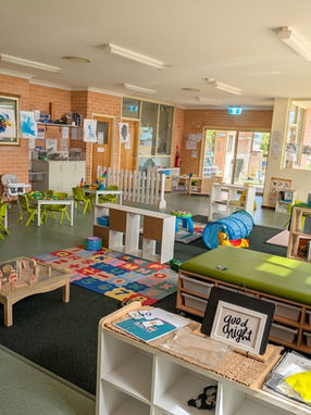 A wide-angle view of the spacious indoor early learning classroom at Clementson Drive Early Educational Centre. The image features distinct, interconnected activity zones: a reading area with low shelving and a play tunnel, several small-group tables with green chairs and high chairs for meals or art, a central sensory mat with wooden shapes, and a display of colorful children's art on a brick wall. This thoughtfully organized space caters to diverse child development needs and serves families in Leppington, Kemps Creek, Austral, and Bradfield.