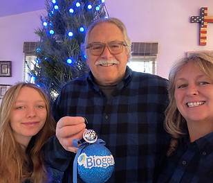 Veteran Don Sladek is standing in front of his Christmas tree with blue lights and holding a blue ornament with the words, "Biogen." His granddaughter, Gracie Sladek is to the left, and to the right is his wife, Chavonne Sladek.