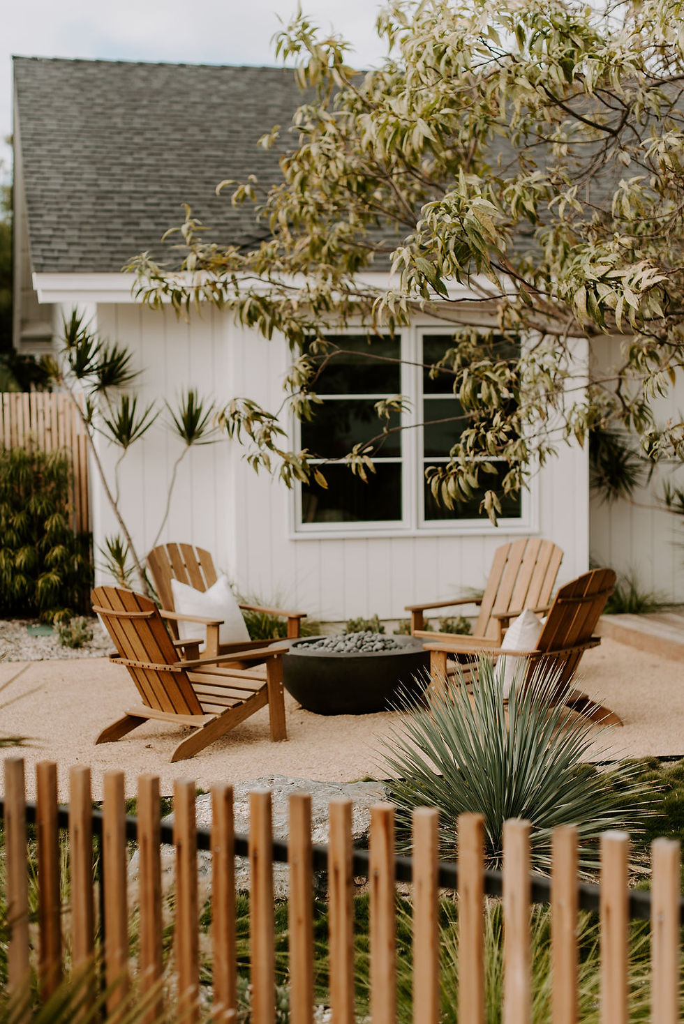 Cozy frontyard patio with four wooden chairs around a black fire pit, white house, leafy tree branches, and a wood fence.