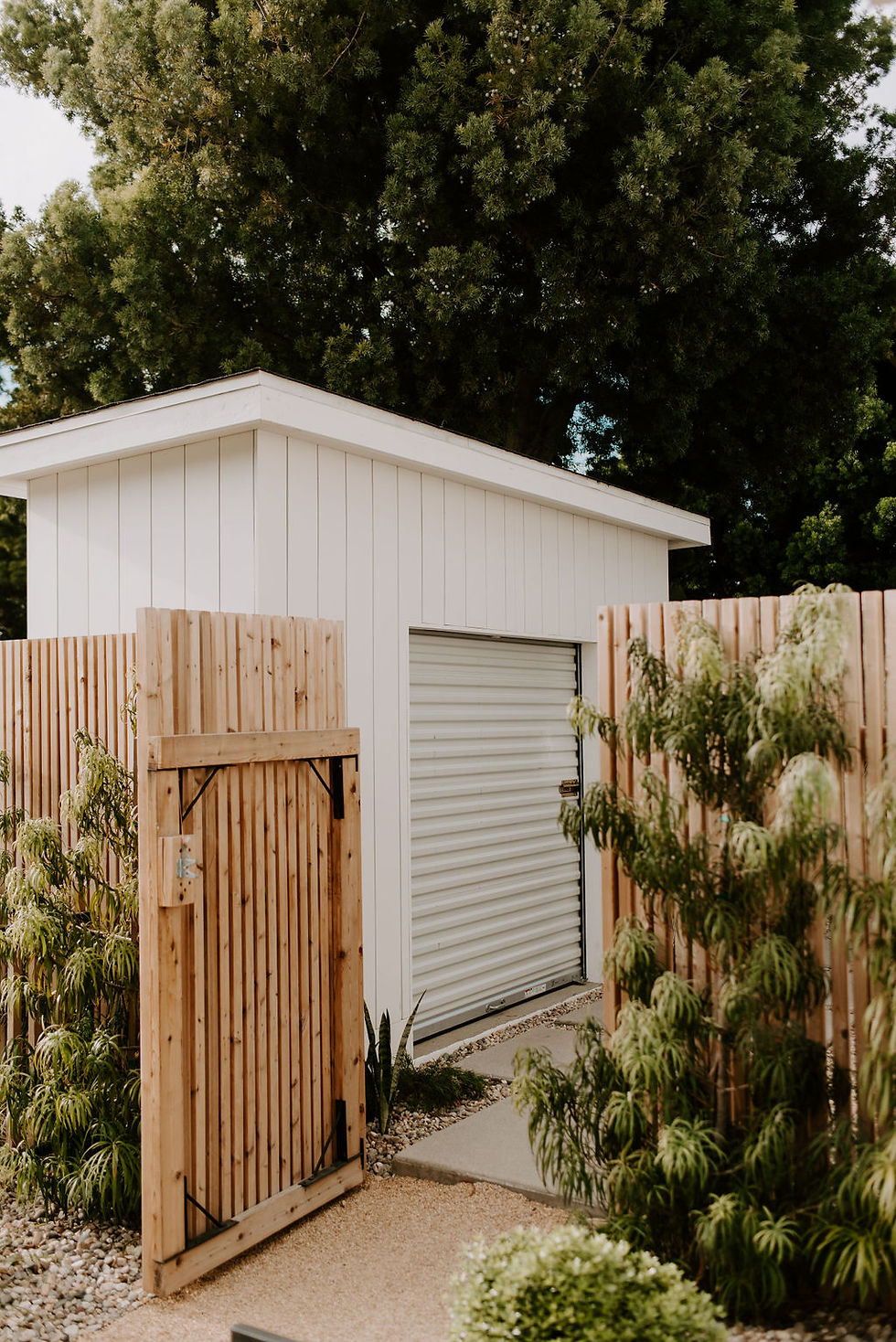 A modern vertical cedar gate opened to reveal a white shed with vertical siding and roll up door.