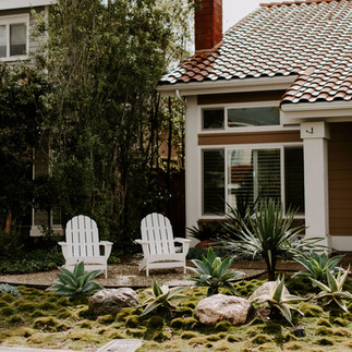 Two white Adirondack chairs sit in a garden with green plants and rocks, near a house with a tiled roof and large windows. Sunny atmosphere.