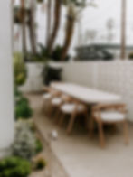 Outdoor dining area with a long oval table, six wooden chairs, and lush plants. Palm trees and foggy sky in the background. Serene mood.