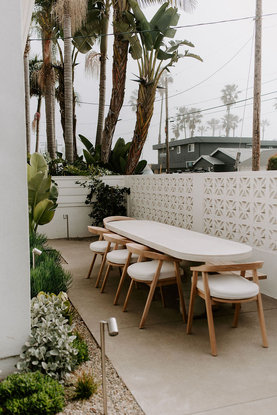 A modern outdoor dining table with teak chairs and a planter bed filled with coastal style plants.