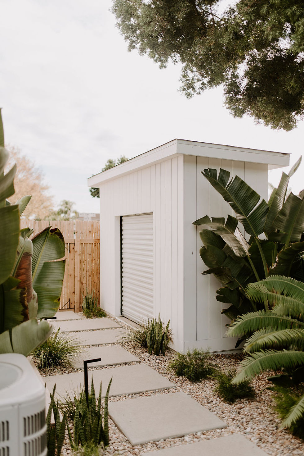 White shed with vertical siding and roll up door flanked with lush tropical landscaping.