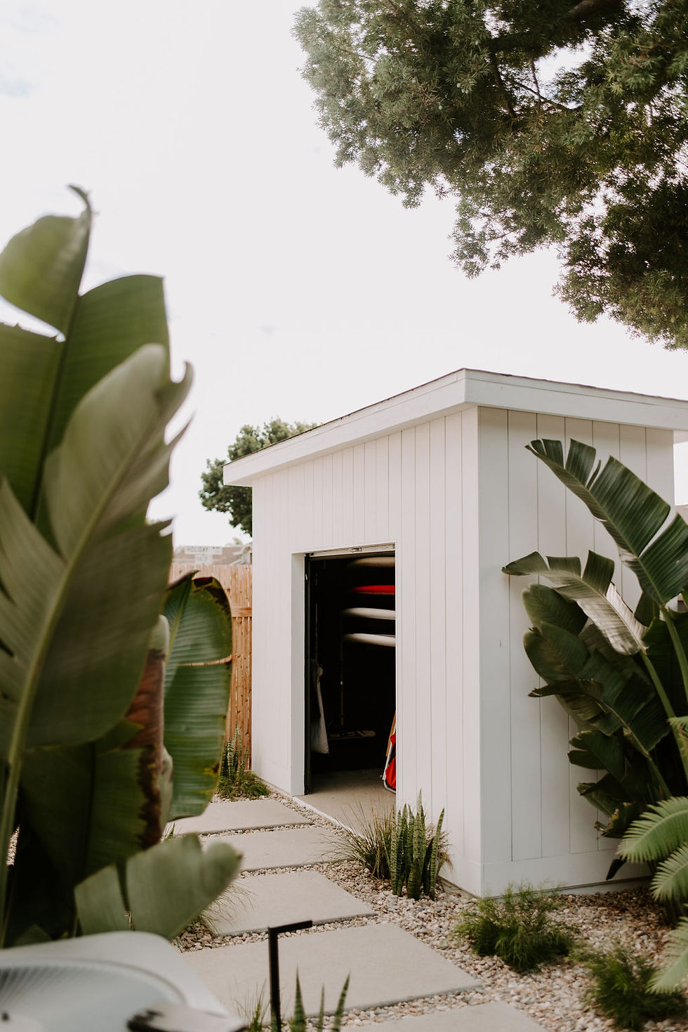 White shed with vertical siding, lush tropical landscaping and stacked surfboards inside.