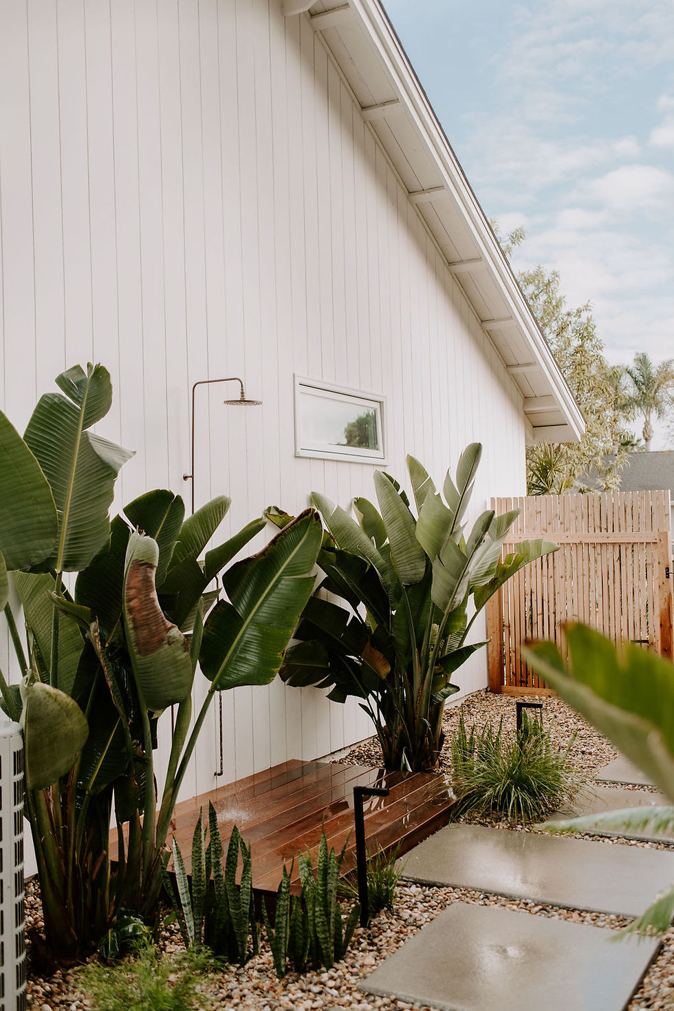 Outdoor shower with wood deck surrounded by palm trees against a white coastal house.