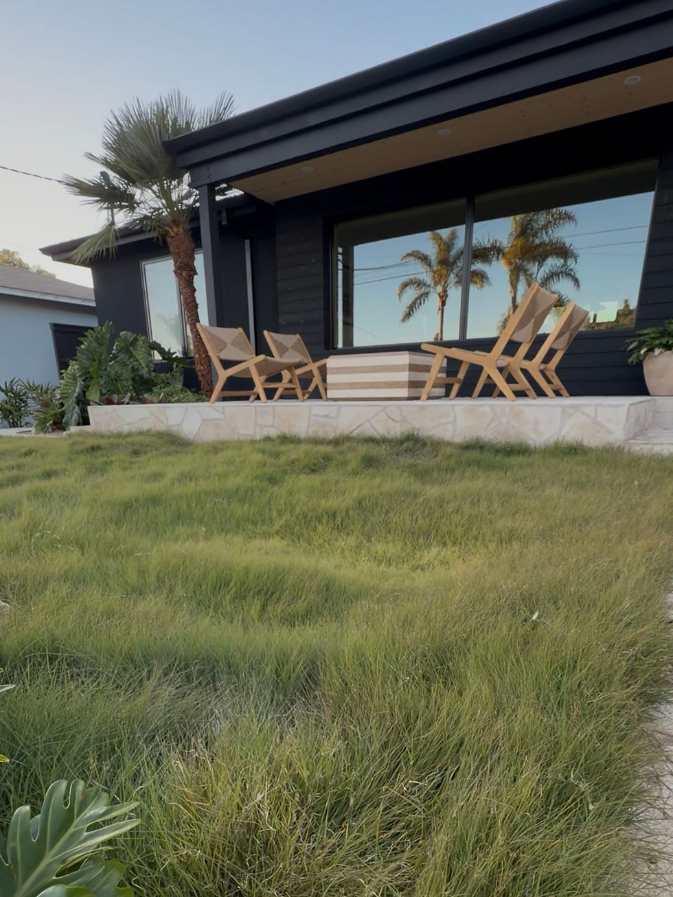 Modern house with black exterior, wooden chairs, and a striped table on the patio. Lush green grass and palm trees in the setting.