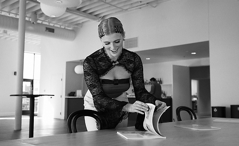 Black and white photo of 1 female flipping through a book on a table. Behind her is a drink bar and table.