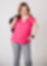 Woman standing looking at camera at a studio for a photo shoot. Wearing bright pink shirt and grey pants. She is holding a black jacket over one of her shoulders.