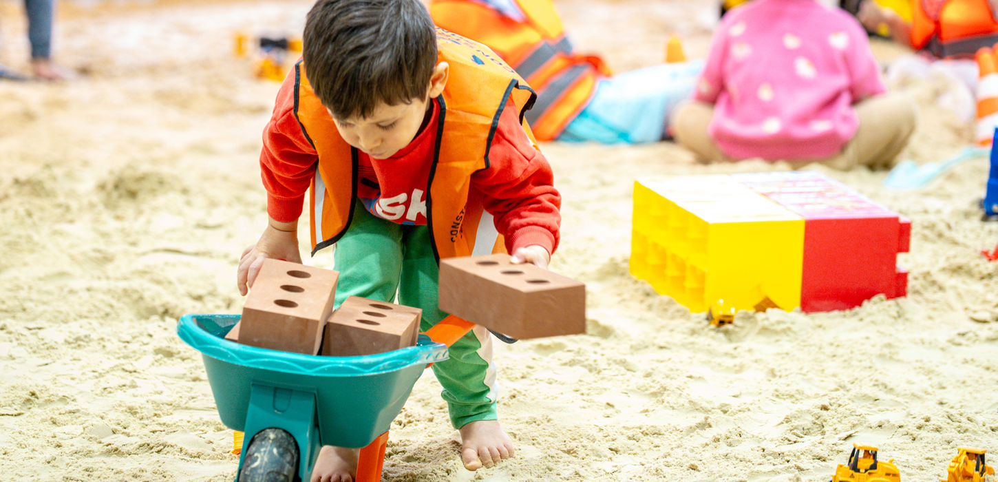 Child in safety vest playing with toy bricks and wheelbarrow