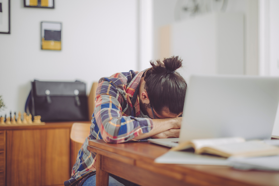 Tired person in a plaid shirt rests their head on folded arms at a desk beside a laptop in a home office.