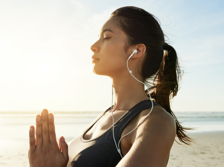 Woman meditating on a beach at sunset, eyes closed, wearing earphones and a tank top. Calm and serene mood with a glowing sun backdrop.
