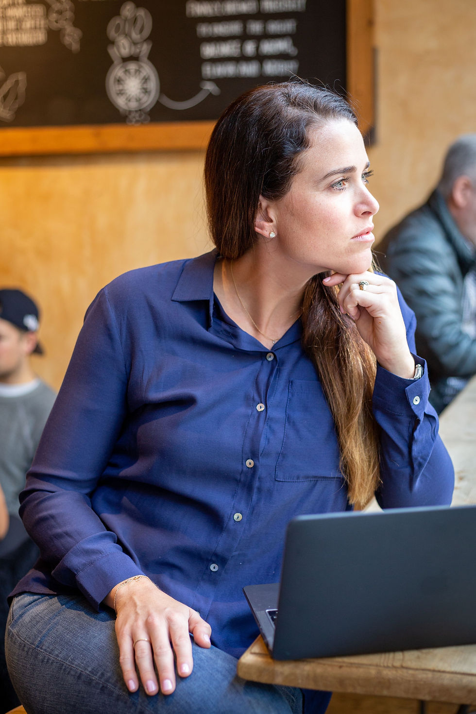 Brooke is in a blue shirt, sitting at a table with a laptop, gazes thoughtfully to the side in a café. Chalkboard menu in background.