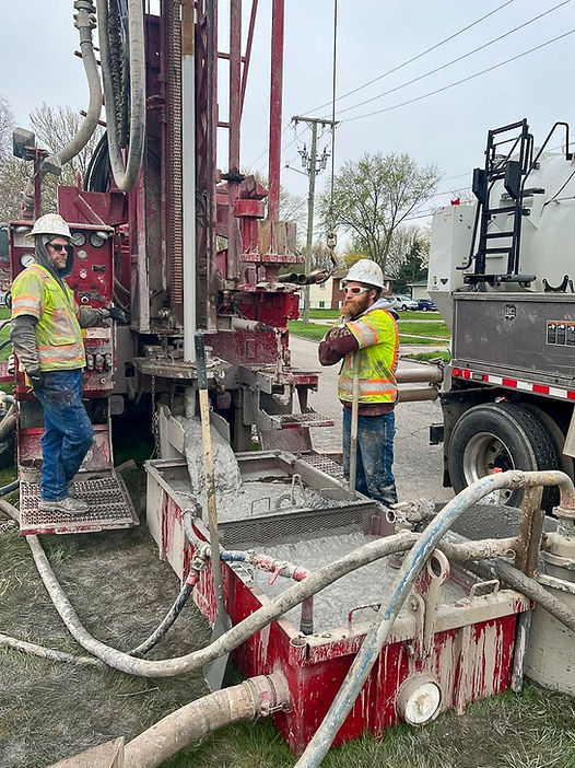 Two workers in high visibility vests operating a large well drilling rig, managing complex machinery and hoses on a residential street.