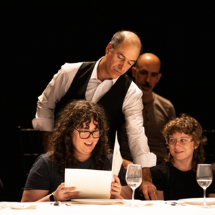 Geoff Sobelle looks of the shoulder of a woman reading a menu at a dinner table. Others sit nearby, set against a dark background.
