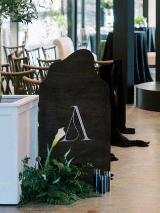 Large-scale organic-shaped welcome sign in matte black with a minimalist white monogram, styled with dark greenery at a modern venue entrance.