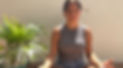Woman meditating outdoors in sunlight with eyes closed, wearing gray tank top and denim shorts, sitting cross-legged beside a potted palm plant.