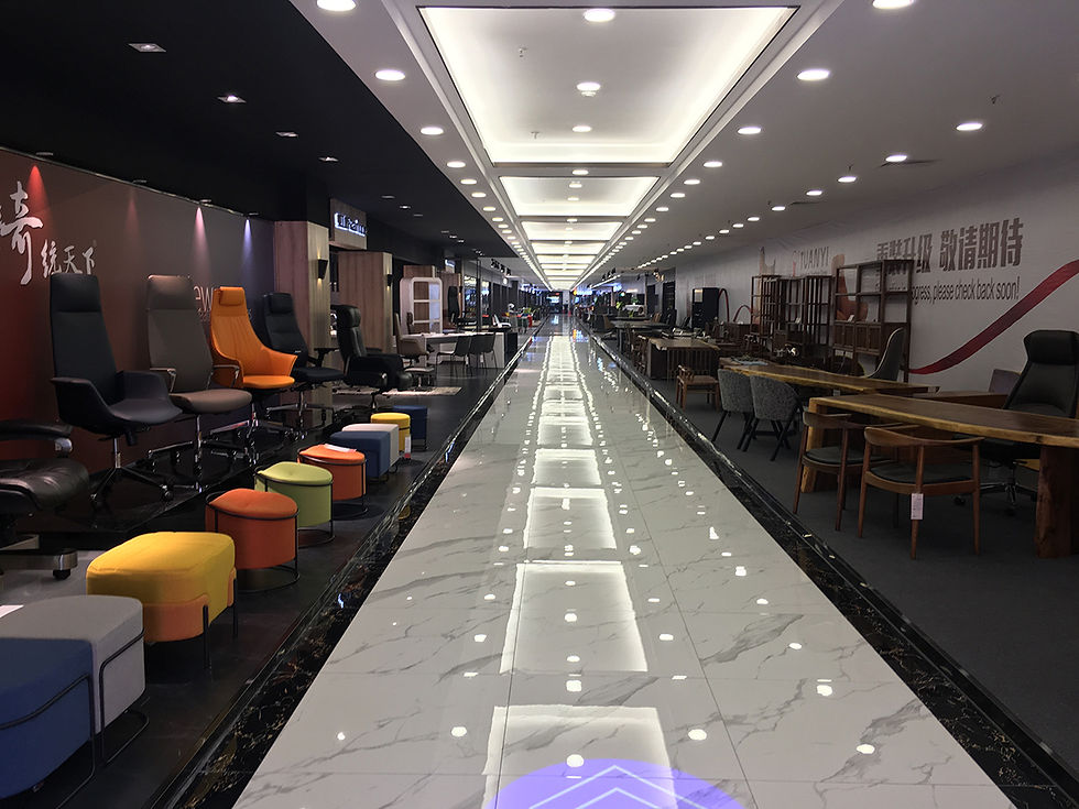 Tuanyi is like a calm, refined cousin of Lecong. It’s compact, organized, and full of practical yet elegant furniture collections.
You’ll find mid-to-premium quality sofas, dining sets, and bedroom furniture with a clean, export-ready finish. Long corridor of a showroom with colorful chairs and tables on either side. Bright overhead lights reflect on the shiny marble floor.